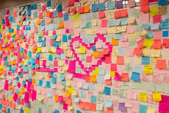 A colorful wall covered in hundreds of sticky notes in pink, blue, yellow, and orange, each with handwritten messages of positivity and gratitude. The notes form a large heart shape in the center, creating a vibrant and inspiring display.