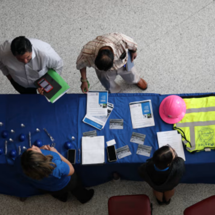 Overhead view of a job fair table with recruiters and attendees reviewing flyers and forms, with a pink hard hat and high-visibility safety vest displayed to represent skilled trade and construction jobs.