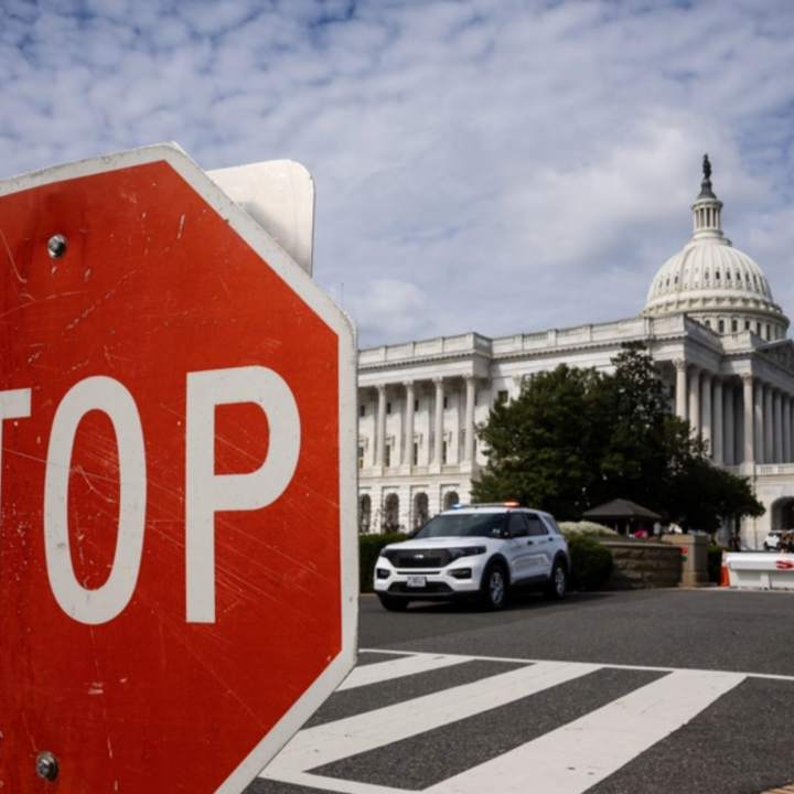 A red stop sign stands in the foreground near a roadblock, with a police vehicle parked nearby and the U.S. Capitol building visible in the background under a partly cloudy sky.