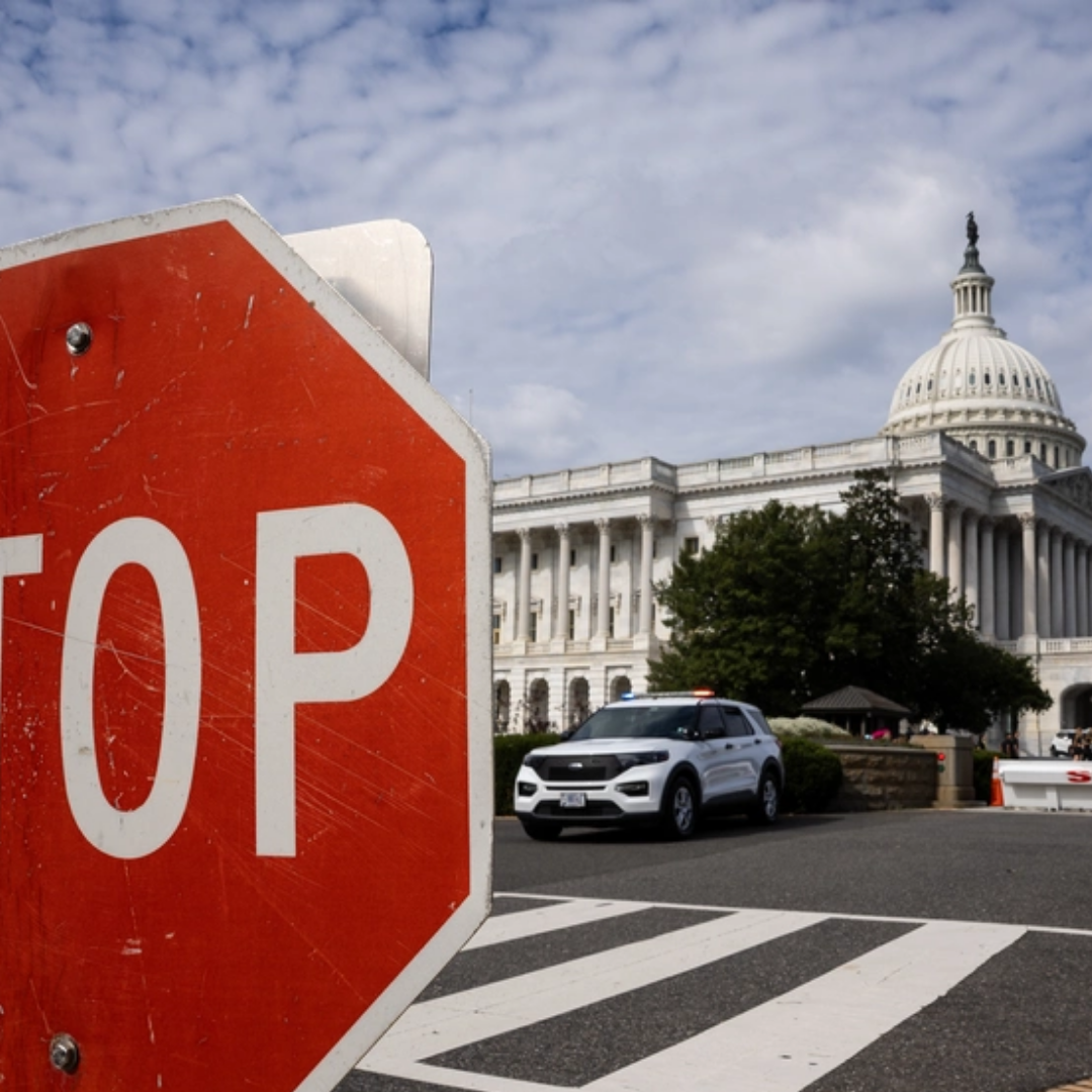 A red stop sign stands in the foreground near a roadblock, with a police vehicle parked nearby and the U.S. Capitol building visible in the background under a partly cloudy sky.