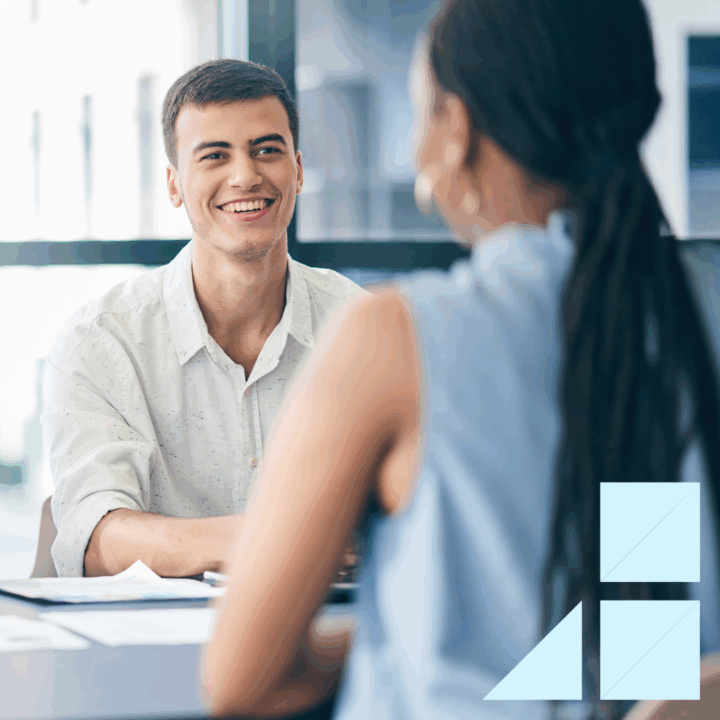 A hiring manager smiles while interviewing a candidate across the table in a modern office. Both are engaged in conversation with documents on the desk, symbolizing a positive and professional hiring decision process.