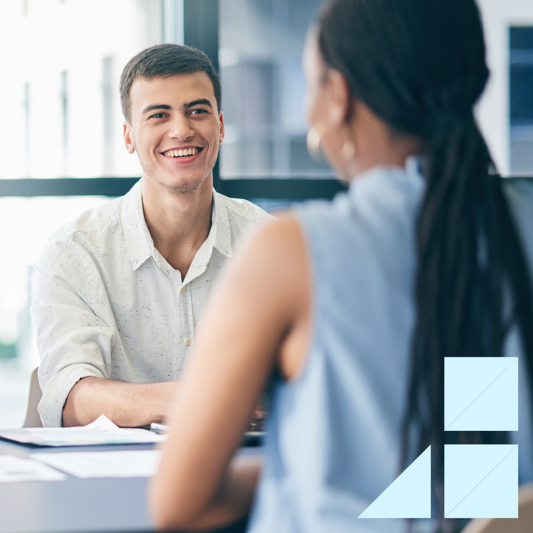 A hiring manager smiles while interviewing a candidate across the table in a modern office. Both are engaged in conversation with documents on the desk, symbolizing a positive and professional hiring decision process.