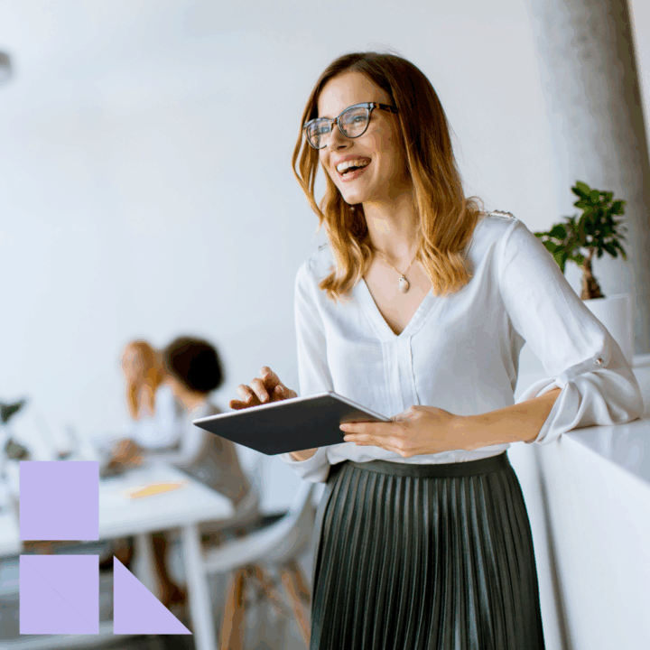 Smiling office manager in a white blouse and pleated skirt holding a tablet while standing in a bright, modern office. Two colleagues collaborate in the background near a desk with laptops, and a small plant adds a touch of greenery to the space.