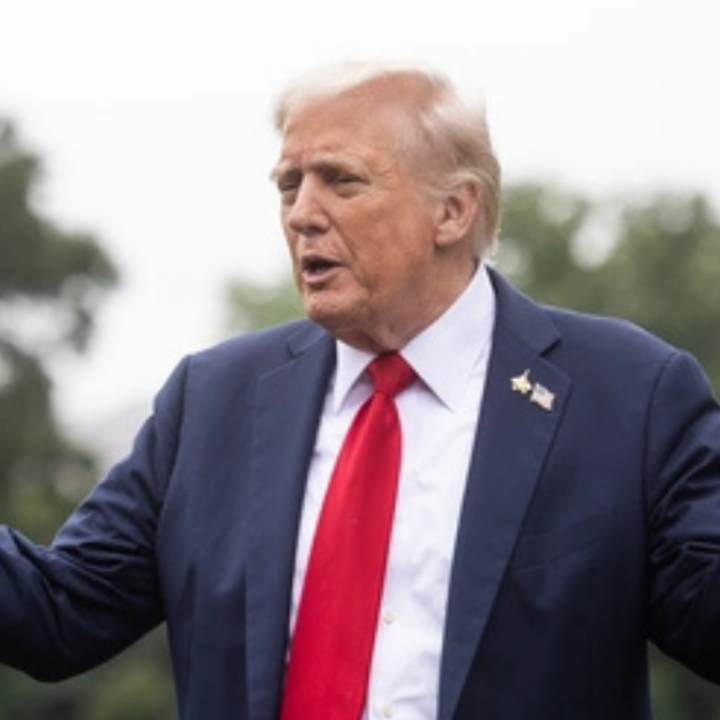 President Trump in a dark suit and red tie gestures with both hands while speaking outdoors, with trees blurred in the background.