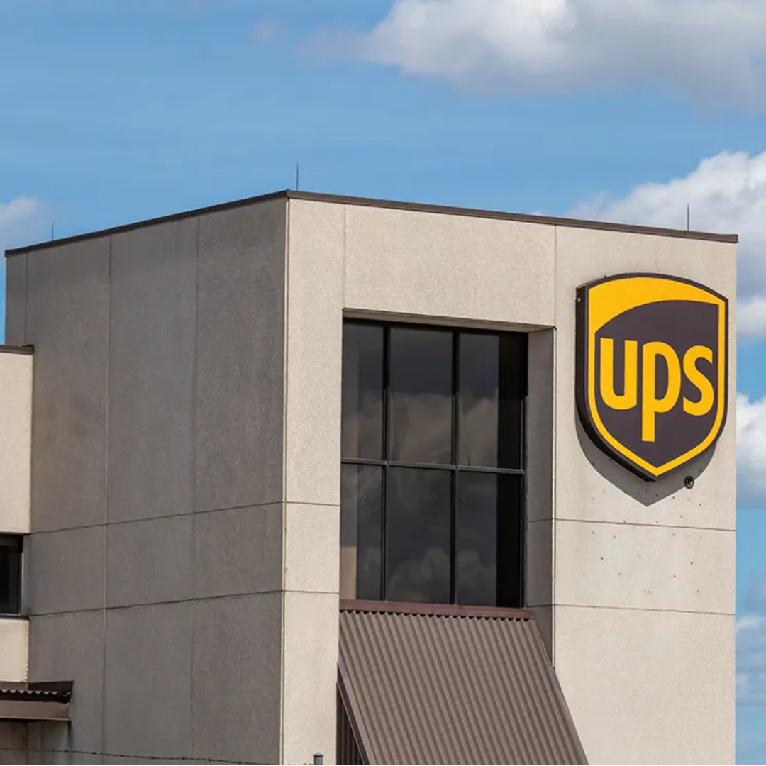 The exterior of a UPS office building is shown, featuring the UPS logo mounted on the building against a blue sky with scattered clouds.