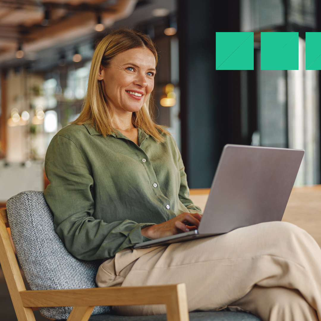 A smiling woman sits in a modern office lounge area, working on a laptop looting at a top applicant tracking system. She wears a light green blouse and beige pants, appearing relaxed and focused. The background shows a bright, open workspace with natural light and contemporary decor.