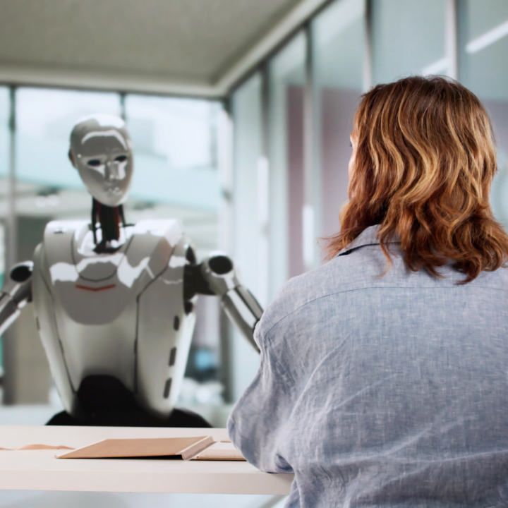 A person sitting at a desk faces a humanoid robot across a table in a modern office setting, suggesting a job interview or hiring discussion involving artificial intelligence.