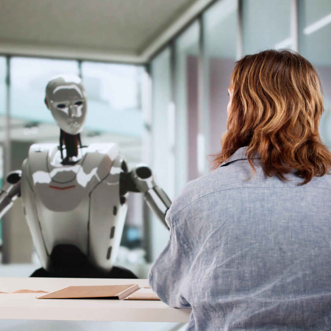A person sitting at a desk faces a humanoid robot across a table in a modern office setting, suggesting a job interview or hiring discussion involving artificial intelligence.