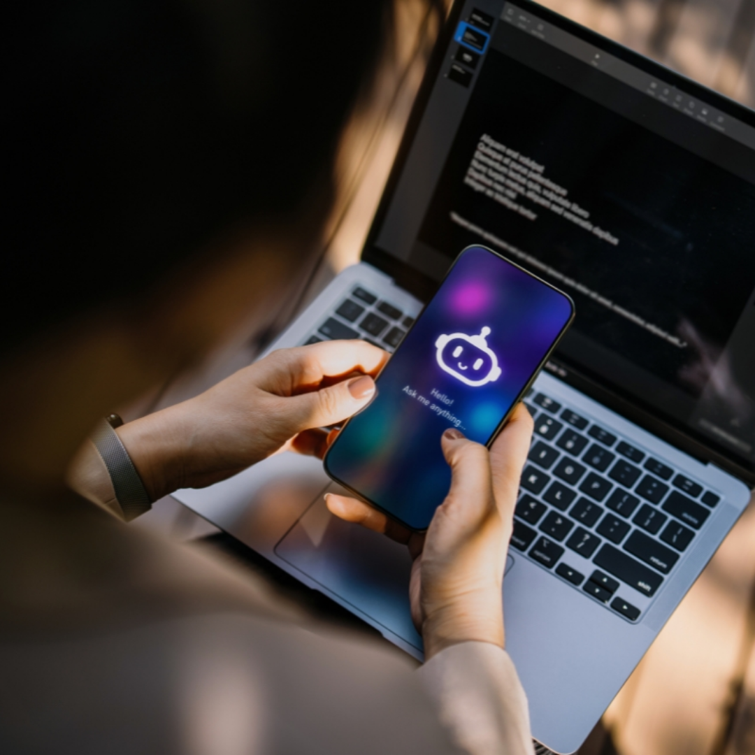 A person holds a smartphone displaying a chatbot interface while sitting at a laptop with code or text visible on the screen, suggesting the use of AI technology alongside computer work.