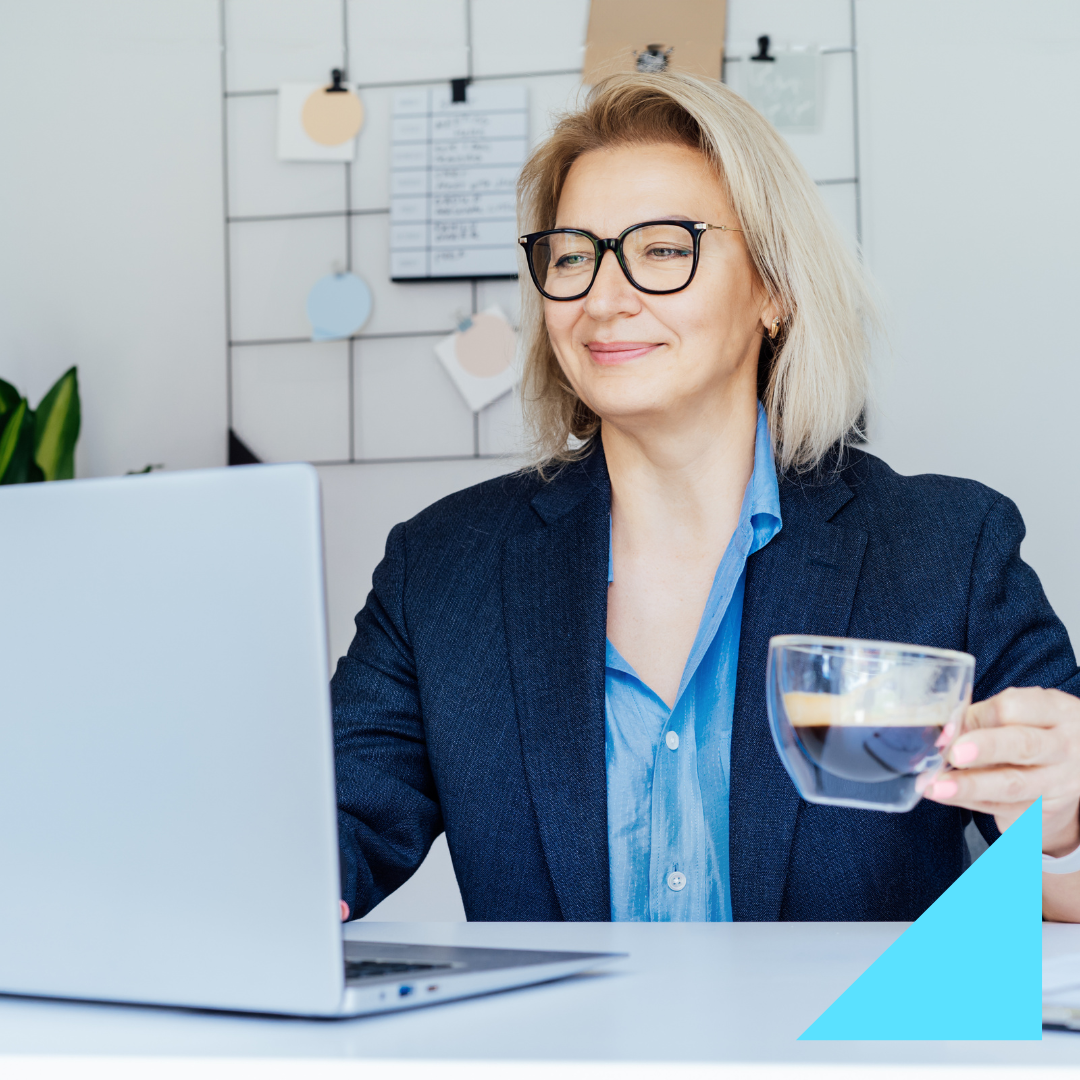 Professional woman with blonde hair and glasses smiling while analyzing source of hire data on her laptop, holding a cup of coffee in a bright modern office with a wall organizer and notes in the background.
