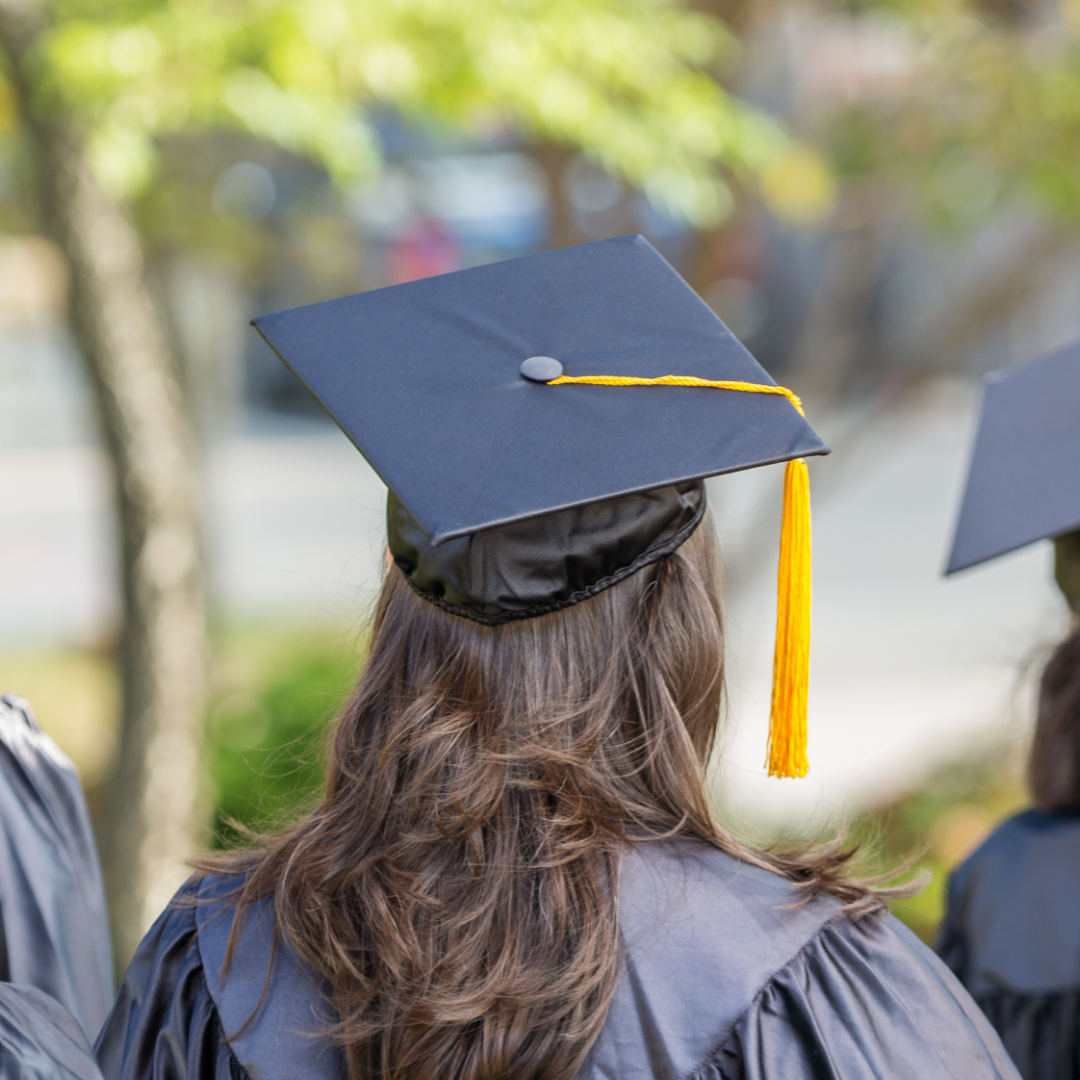 A college graduate wearing a black cap and gown is seen from behind during an outdoor graduation ceremony, with trees softly blurred in the background.