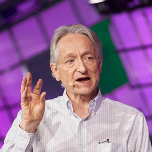 Older man speaking on stage with a headset microphone, gesturing with one hand against a purple and green geometric backdrop.