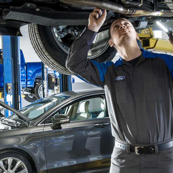 An auto mechanic inspects the underside of a car raised on a hydraulic lift inside a repair shop, with another vehicle and service bays visible in the background.