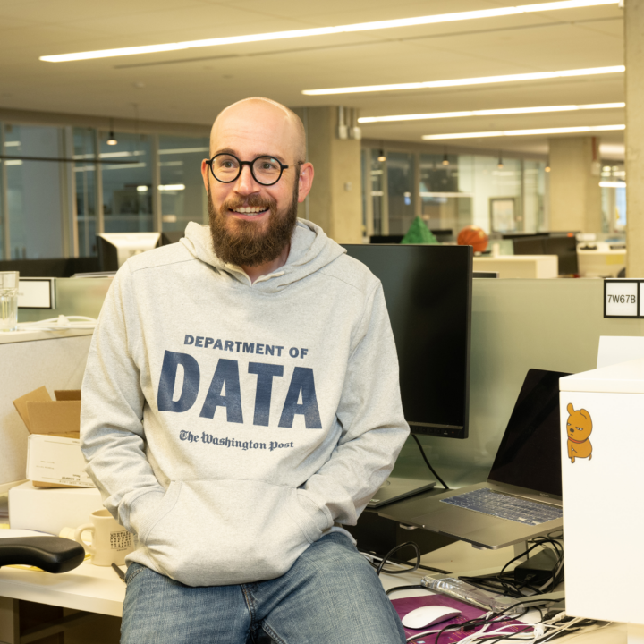 A man wearing glasses and a hoodie that reads “Department of Data” sits at a desk in a modern office, smiling, with computer monitors and work items visible around him.