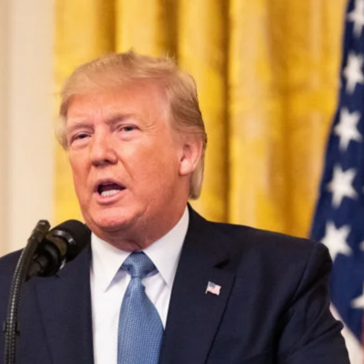President Trump in a dark suit and blue tie speaks into a microphone indoors, with gold curtains and a U.S. flag visible in the background.
