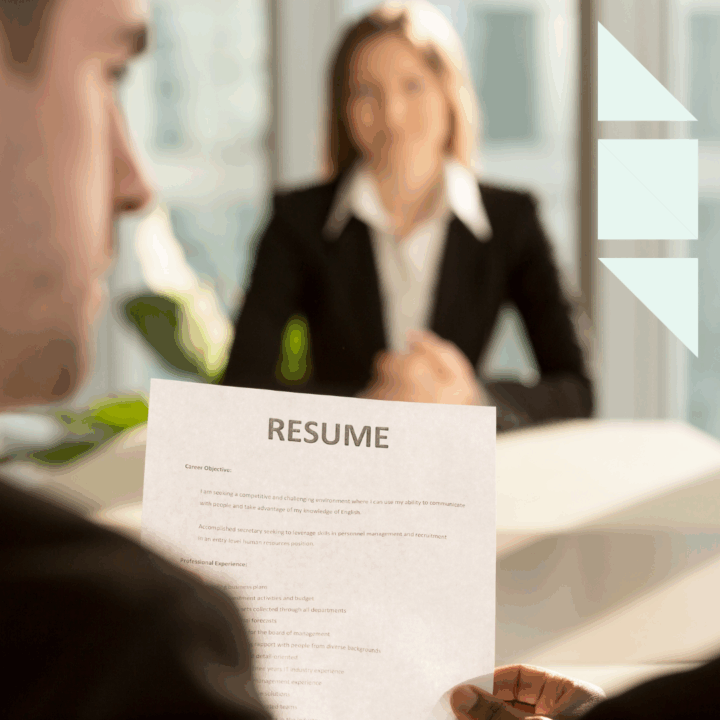 A close-up view of a hiring manager holding and reviewing a printed resume during an in-person job interview. The resume text is partially visible in focus, while a professionally dressed candidate sits across the desk slightly out of focus in the background. The office setting features natural light and a modern, professional atmosphere, emphasizing the concept of candidate evaluation and resume screening.