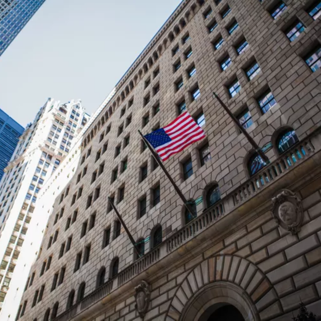 American flag flying outside a large stone office building in a downtown area, viewed from street level with surrounding skyscrapers in the background.