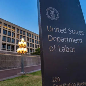 United States Department of Labor sign outside a federal office building at dusk, with the agency seal visible and office windows illuminated in the background.