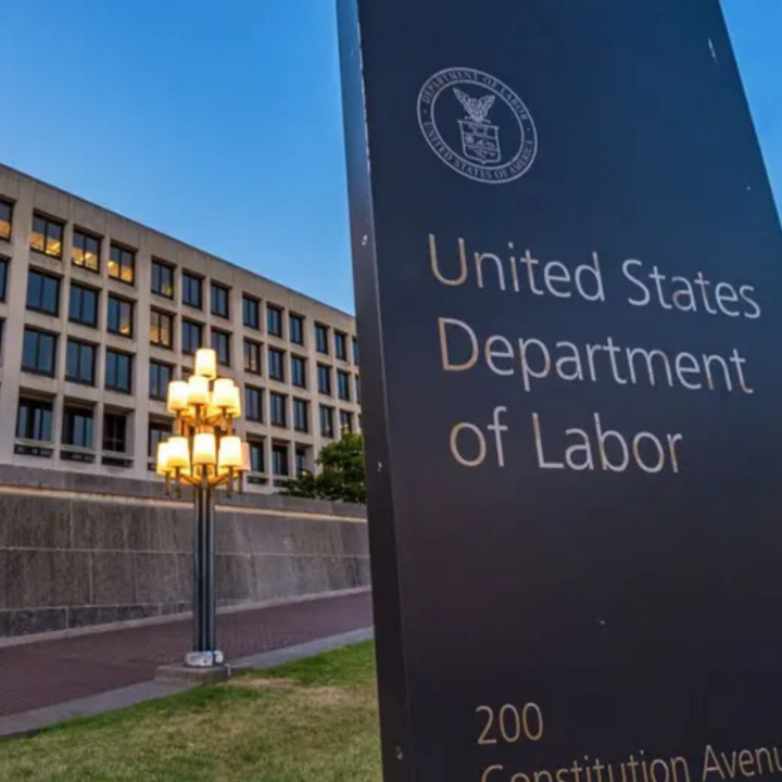 United States Department of Labor sign outside a federal office building at dusk, with the agency seal visible and office windows illuminated in the background.