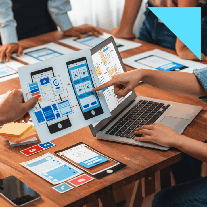 A team of UX designers reviewing mobile app wireframes and prototypes at a table, with one person pointing at layout designs on a laptop screen.