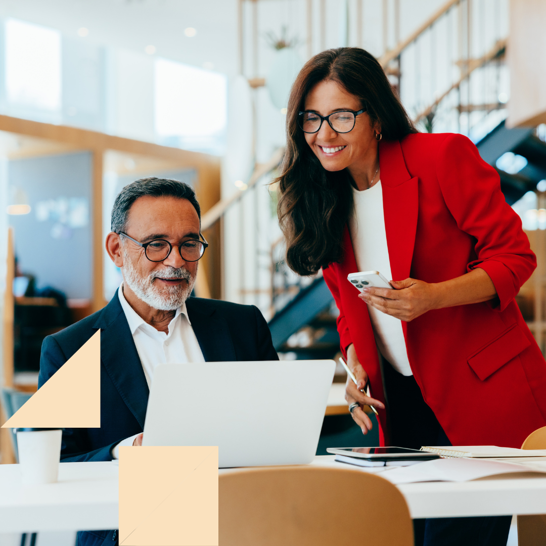 A small business recruiter collaborates with a hiring manager in a modern office, reviewing candidates on a laptop as they discuss staffing needs; the recruiter stands beside him smiling while holding a phone, demonstrating a friendly, professional hiring partnership.