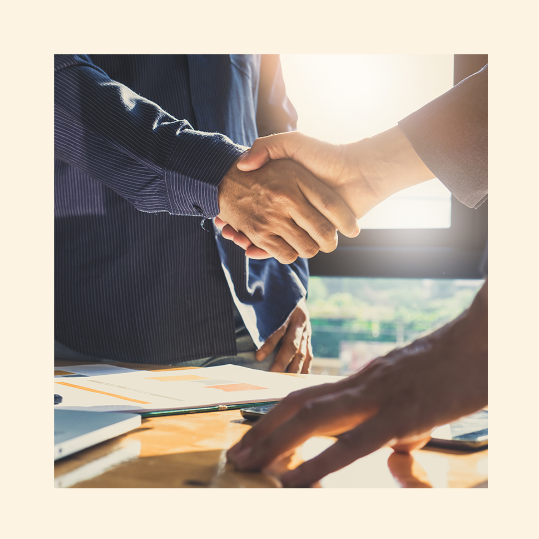 HR manager and third-party recruiter shaking hands across a desk during a job offer or interview meeting, with documents and a laptop visible in a professional office setting.