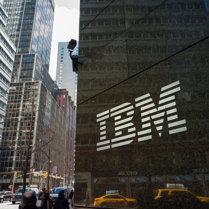 IBM logo displayed on the exterior of a modern office building in an urban setting, with city streets and skyscrapers visible in the background.