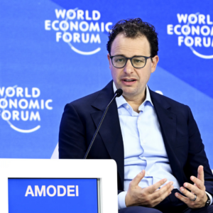 Man speaking at a panel discussion with a microphone, seated in front of a blue backdrop displaying “World Economic Forum” logos, wearing glasses and a dark blazer over a light shirt.
