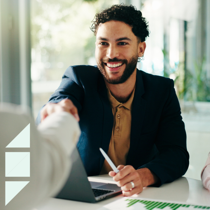 Smiling candidate in a business-casual blazer shaking hands across a desk during a tech interview, with a laptop, stylus, and interview materials visible in a bright modern office.