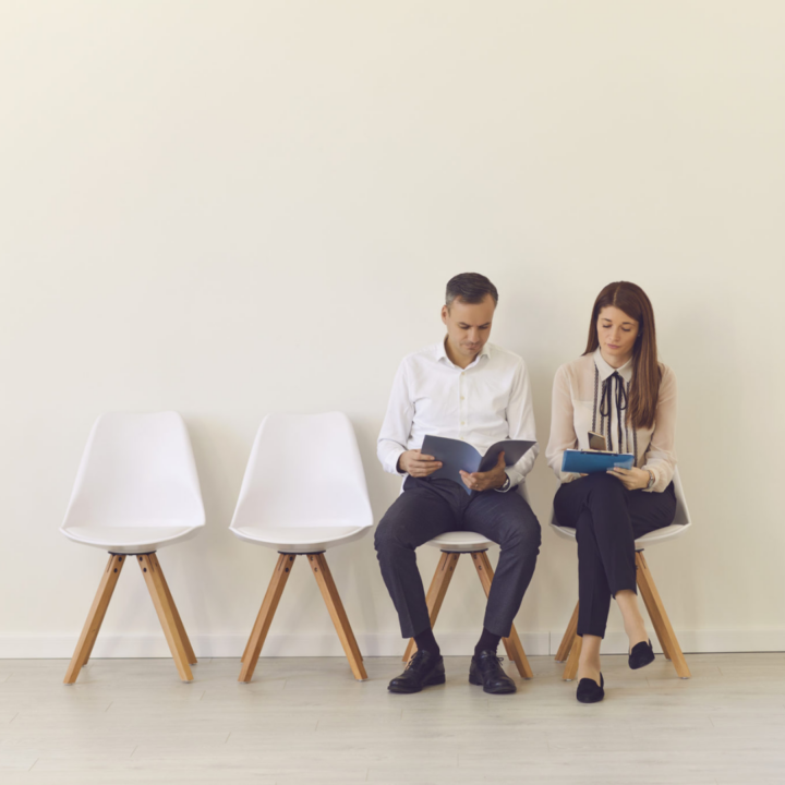 Two job seekers seated in a waiting area reviewing documents, with empty chairs beside them, suggesting preparation for interviews or employment opportunities.