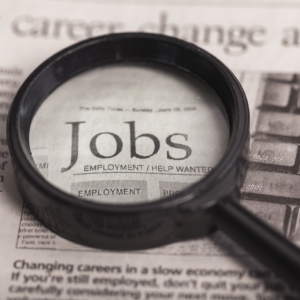 Alt text: Magnifying glass placed over a newspaper classified section highlighting the word “Jobs,” representing job searching or employment opportunities.