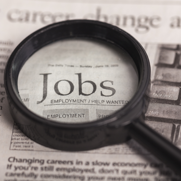 Alt text: Magnifying glass placed over a newspaper classified section highlighting the word “Jobs,” representing job searching or employment opportunities.