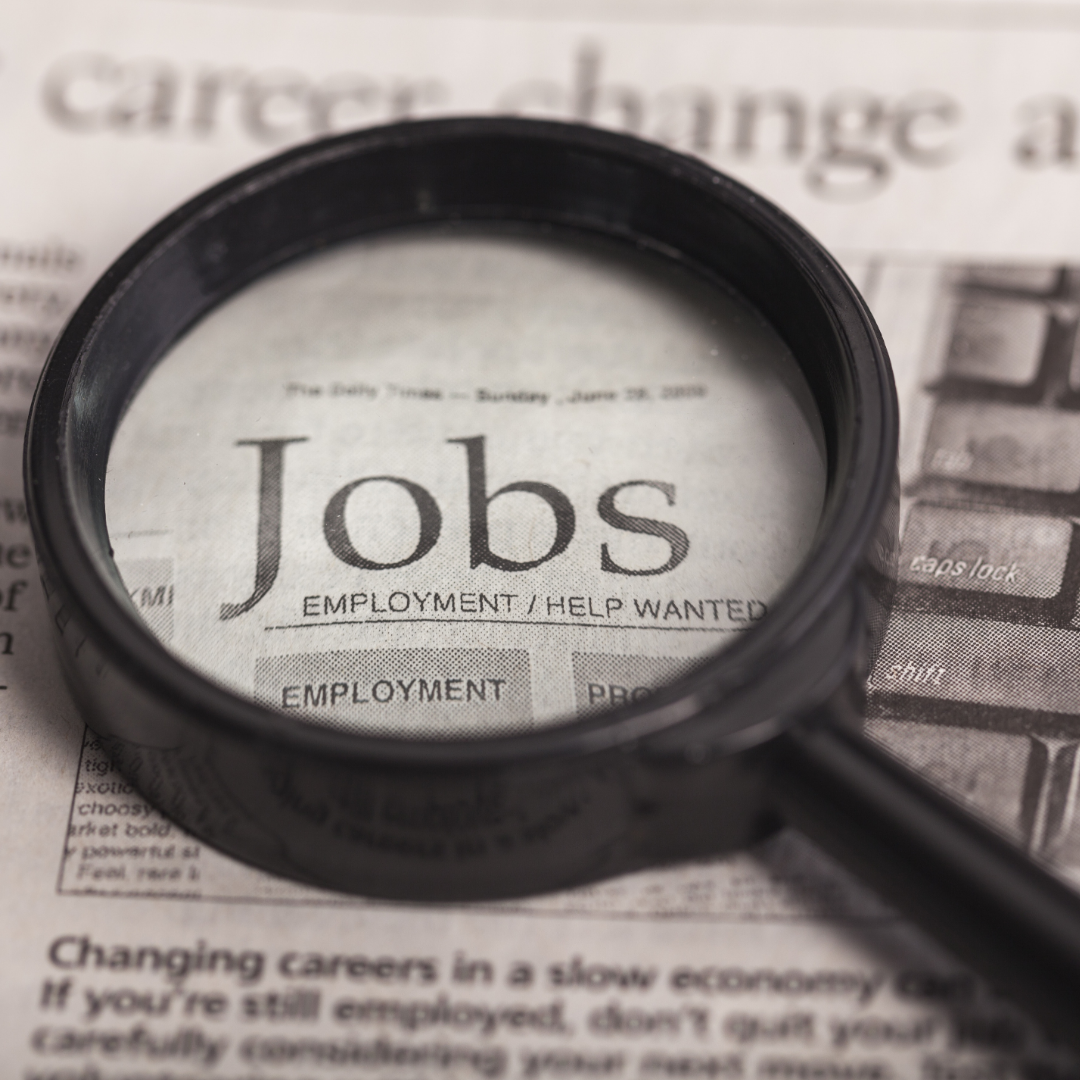 Alt text: Magnifying glass placed over a newspaper classified section highlighting the word “Jobs,” representing job searching or employment opportunities.
