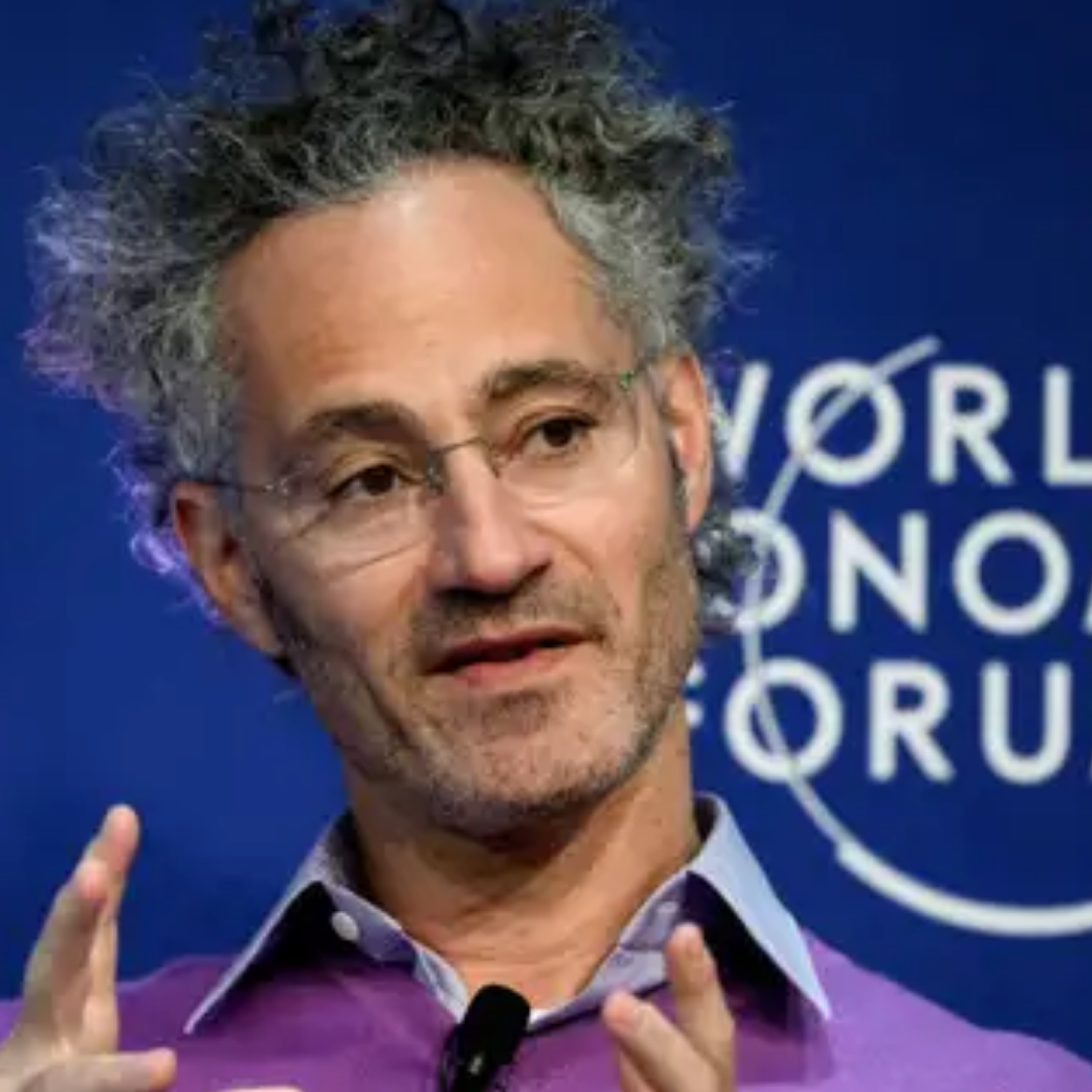 Man speaking at a World Economic Forum event, wearing glasses and a purple sweater, gesturing with his hands in front of a blue backdrop with forum branding.