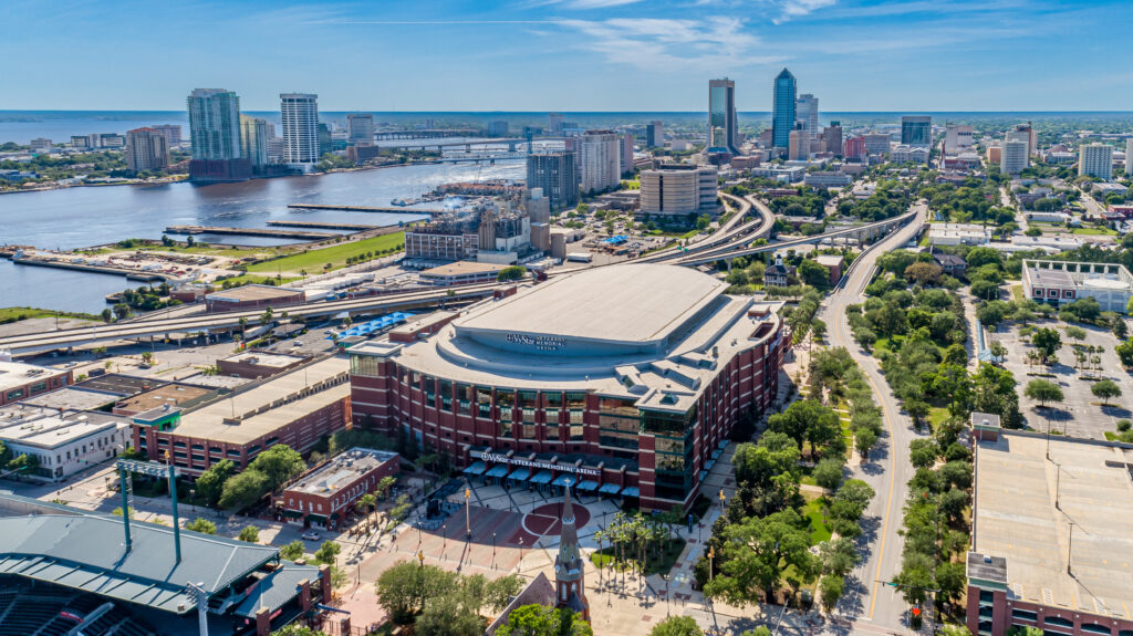 Aerial view of downtown Jacksonville, Florida featuring VyStar Veterans Memorial Arena in the foreground, the St. Johns River, city skyline, bridges, and surrounding highways on a clear day.