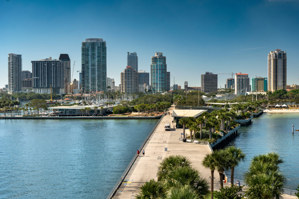 Waterfront view of downtown St. Petersburg, Florida featuring a long pier lined with palm trees extending into Tampa Bay, with modern high-rise buildings and marinas under a clear blue sky.