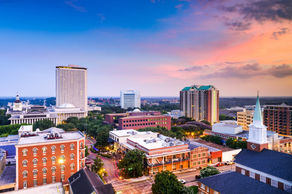 Aerial view of downtown Tallahassee, Florida at sunset, featuring the Florida State Capitol building, historic red-brick buildings, a church steeple, and high-rise offices under a colorful pink and blue sky.