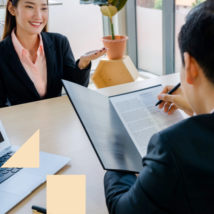 Hiring manager reviewing a candidate’s resume during an in-person interview, holding a clipboard and taking notes while the candidate speaks across a desk with a laptop and plant visible in a modern office setting.