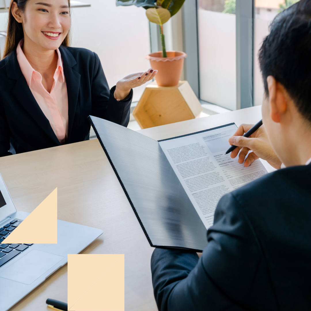 Hiring manager reviewing a candidate’s resume during an in-person interview, holding a clipboard and taking notes while the candidate speaks across a desk with a laptop and plant visible in a modern office setting.