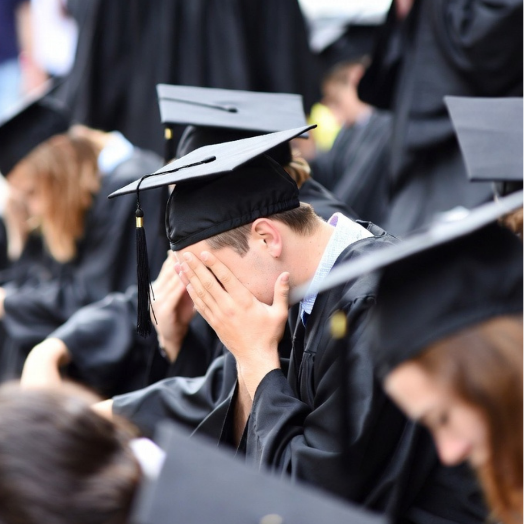 College graduate in cap and gown sitting with head in hands during graduation ceremony