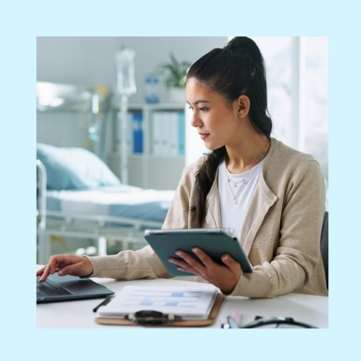Medical billing professional working at desk with laptop and tablet, reviewing healthcare documents in clinical office setting