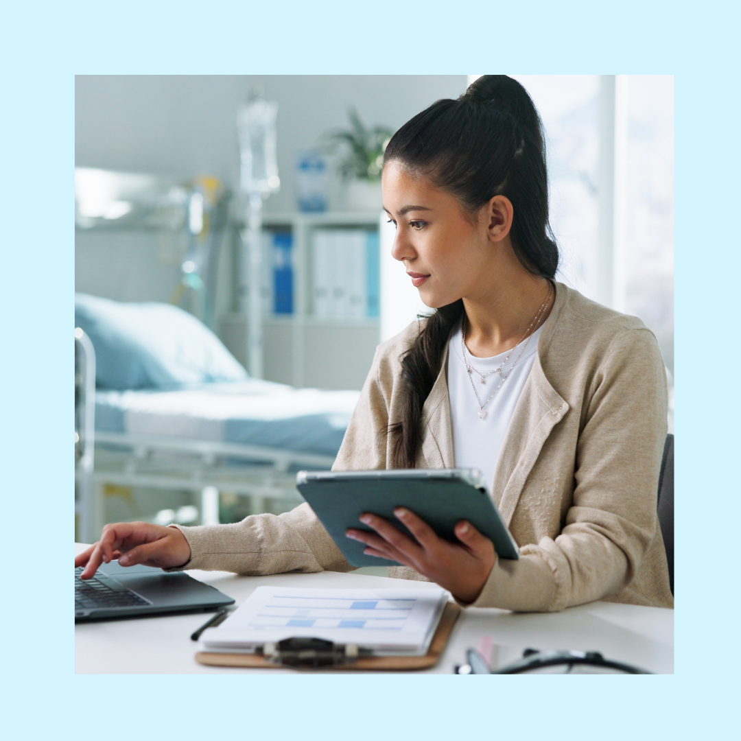 Medical billing professional working at desk with laptop and tablet, reviewing healthcare documents in clinical office setting