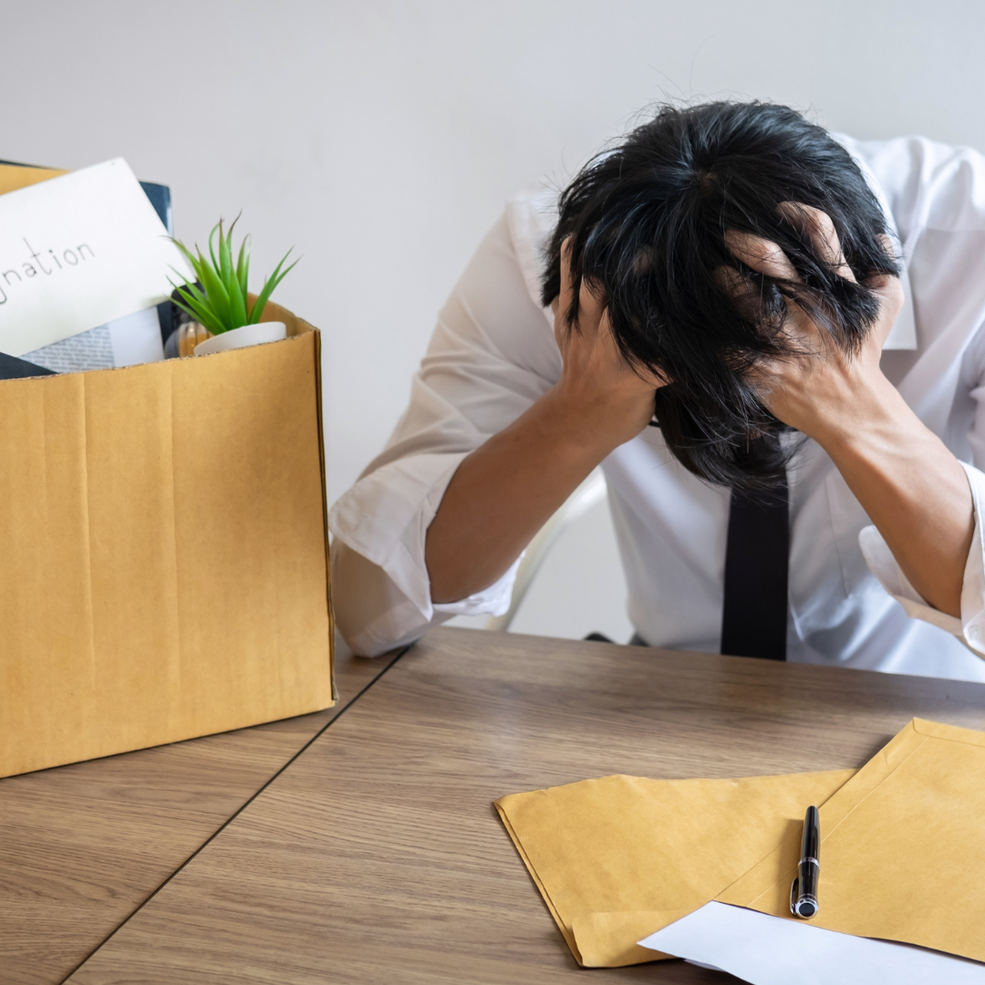 Stressed office worker sitting at a desk with their head in their hands next to a cardboard box of belongings, suggesting job loss, resignation, or a difficult career decision.