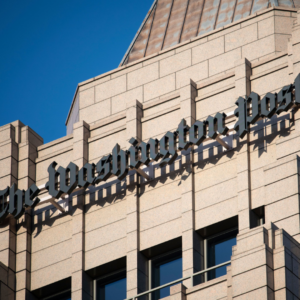 Exterior view of a stone office building with The Washington Post name displayed in black lettering against a clear blue sky.