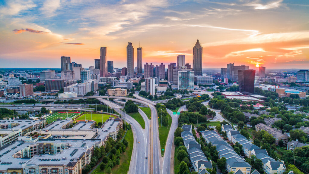 Aerial view of the Atlanta, Georgia skyline at sunset with downtown skyscrapers, intersecting highways, residential neighborhoods, and a football field in the foreground under a colorful evening sky.