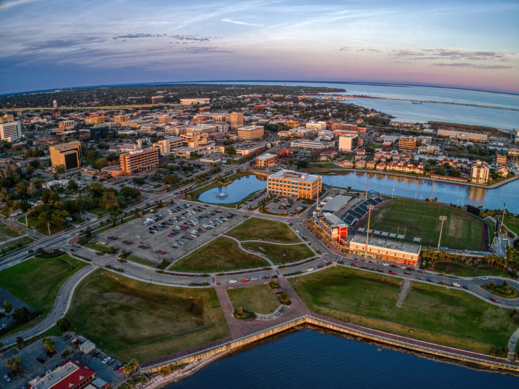 Aerial view of downtown Pensacola, Florida at sunset, featuring a waterfront park, marina, football stadium, and city buildings along Pensacola Bay under a colorful evening sky.
