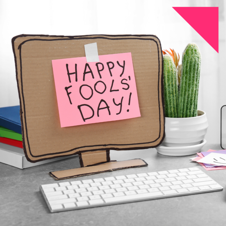 Cardboard computer monitor with a pink sticky note that reads “Happy Fool’s Day!” sitting on an office desk in front of a keyboard, with books and a small cactus plant in the background as part of an April Fools’ Day office prank setup.