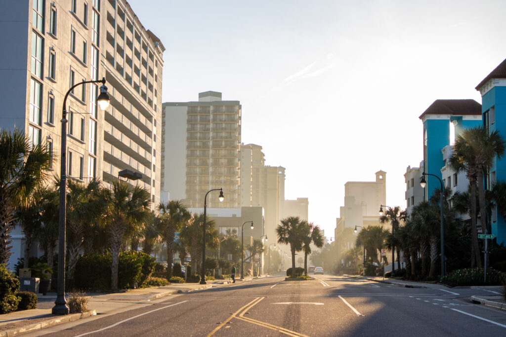 Street view of downtown Myrtle Beach, South Carolina with palm trees, beachfront hotels, and early morning sunlight illuminating the coastal boulevard.