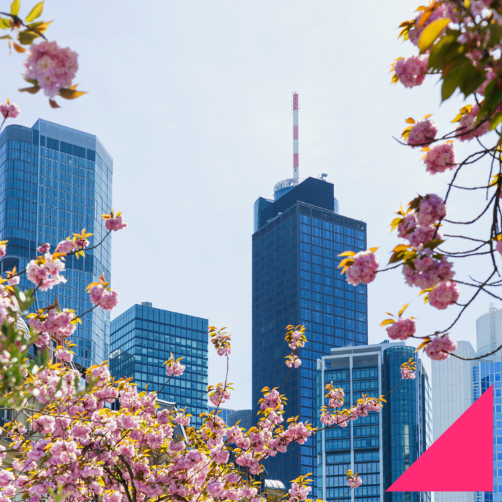 Modern city skyline with blue glass office buildings framed by blooming pink spring flowers, set against a bright hazy sky, with a pink geometric graphic in the bottom-right corner.
