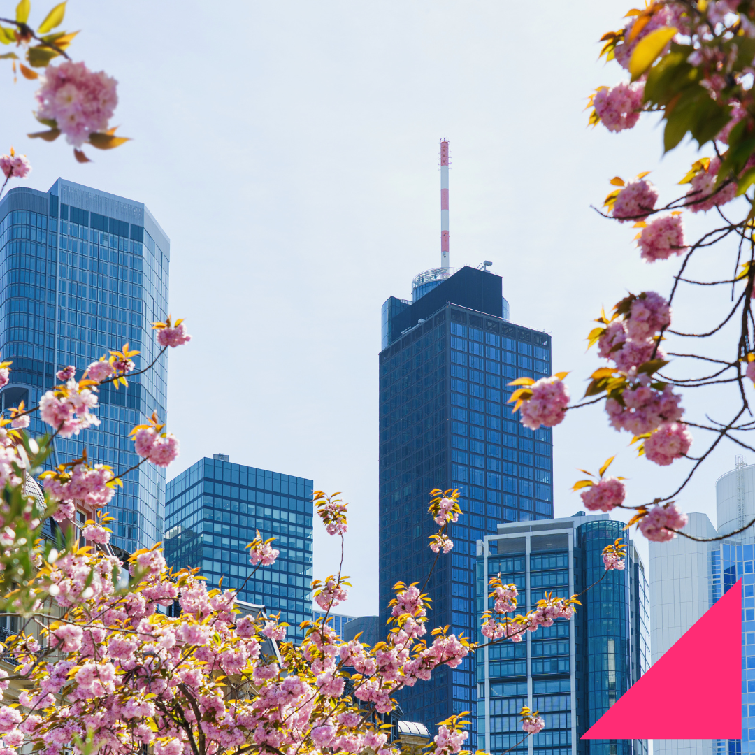 Modern city skyline with blue glass office buildings framed by blooming pink spring flowers, set against a bright hazy sky, with a pink geometric graphic in the bottom-right corner.